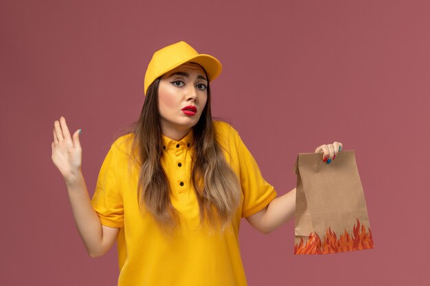 Front view of female courier in yellow uniform and cap holding food package on light-pink wall