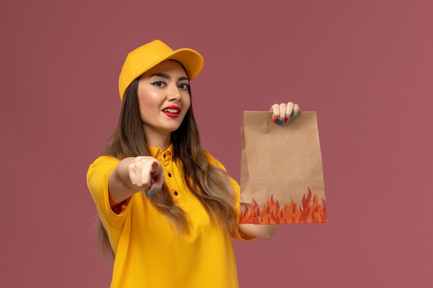 Вид спереди курьера-женщины в желтой форме и кепке, держащей пакет с продуктами на светло-розовой стене