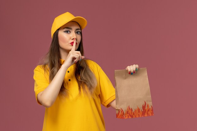 Front view of female courier in yellow uniform and cap holding food package on light-pink wall