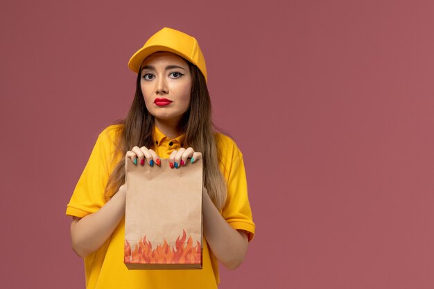 黄色のユニフォームと薄ピンクの壁に食品パッケージを保持しているキャップの女性の宅配便の正面図