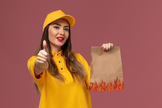 Front view of female courier in yellow uniform and cap holding food package on light-pink wall