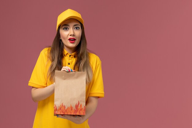 Front view of female courier in yellow uniform and cap holding food package on the light-pink wall