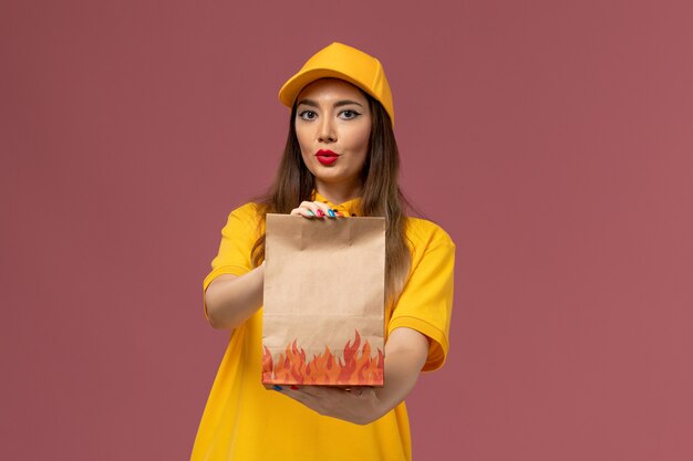 Front view of female courier in yellow uniform and cap holding food package on the light-pink wall