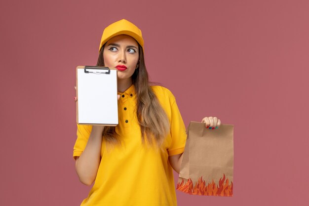 Front view of female courier in yellow uniform and cap holding food package and notepad thinking on the pink wall