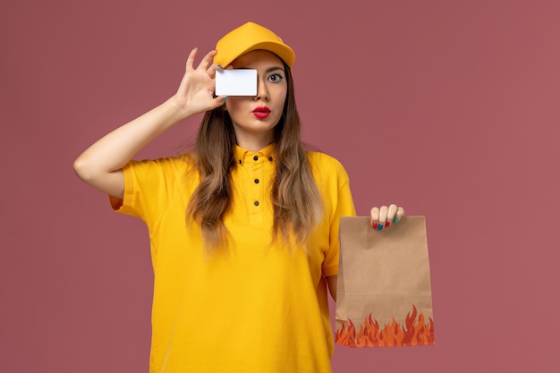 Front view of female courier in yellow uniform and cap holding food package and plastic card on pink wall