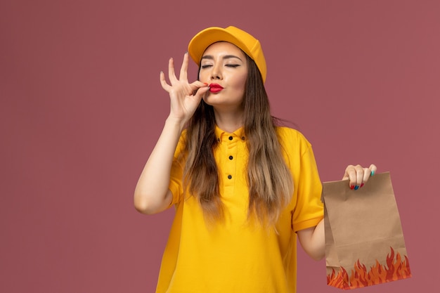 Вид спереди курьера-женщины в желтой форме и кепке, держащей продуктовый пакет со знаком вкуса на светло-розовой стене