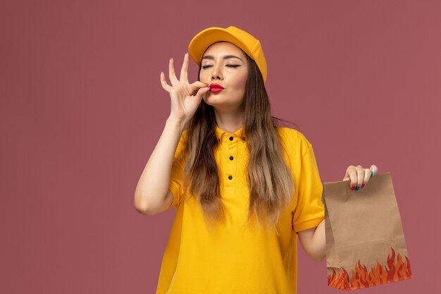 Front view of female courier in yellow uniform and cap holding food package showing taste sign on light-pink wall