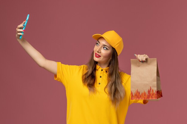 黄色の制服を着た女性の宅配便と食品パッケージを保持し、ピンクの壁で自分撮りをしているキャップの正面図