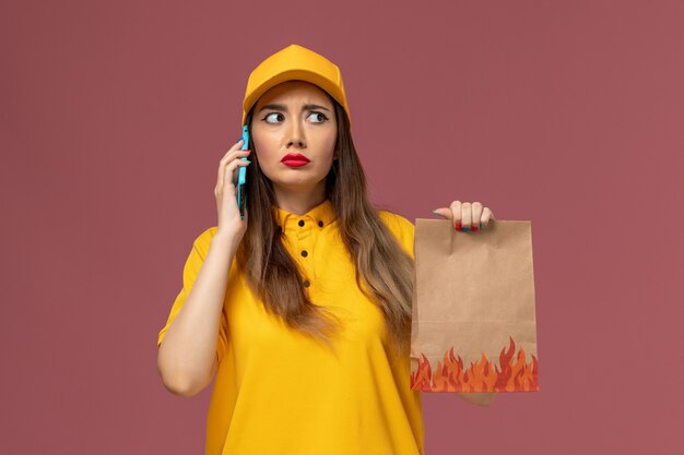 Front view of female courier in yellow uniform and cap holding food package and talking on the phone on pink wall