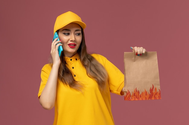 Front view of female courier in yellow uniform and cap holding food package and talking on the phone on pink wall