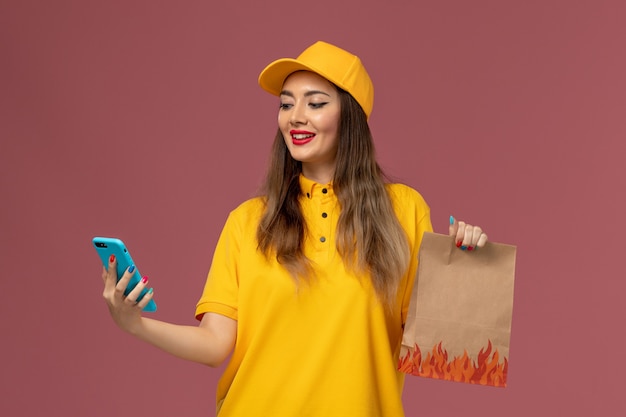 Front view of female courier in yellow uniform and cap holding food package and using a phone on pink wall