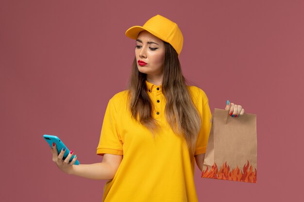 Front view of female courier in yellow uniform and cap holding food package and using a phone on pink wall