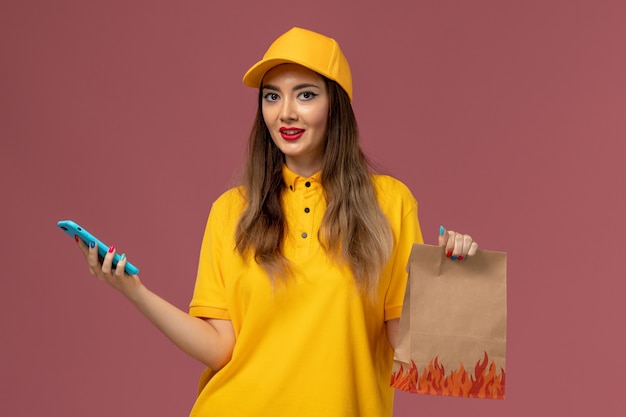 Front view of female courier in yellow uniform and cap holding food package and using a phone on pink wall