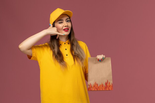 Front view of female courier in yellow uniform and cap holding food package winking on light-pink wall