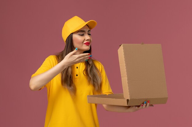 Front view of female courier in yellow uniform and cap holding open food box smelling on light pink wall