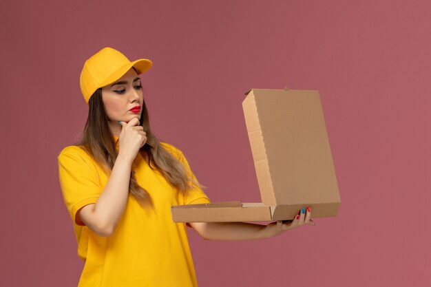 黄色のユニフォームと薄ピンクの壁に考えて開いているフードボックスを保持しているキャップの女性の宅配便の正面