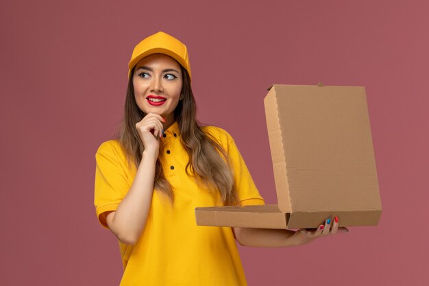 Front view of female courier in yellow uniform and cap holding open food box thinking with smile on light pink wall
