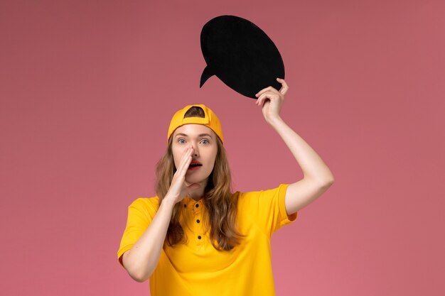 Front view female courier in yellow uniform and cape holding big black sign on pink wall, company service job delivery uniform girl worker