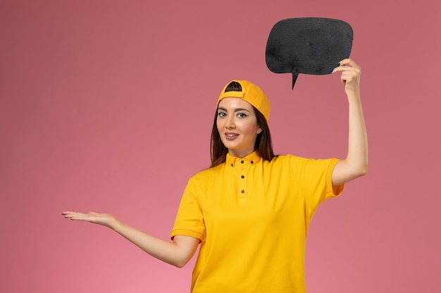 Foto gratuita corriere femminile di vista frontale in uniforme gialla e mantello che tiene grande segno nero sulla parete rosa, consegna del lavoro dell'uniforme di servizio