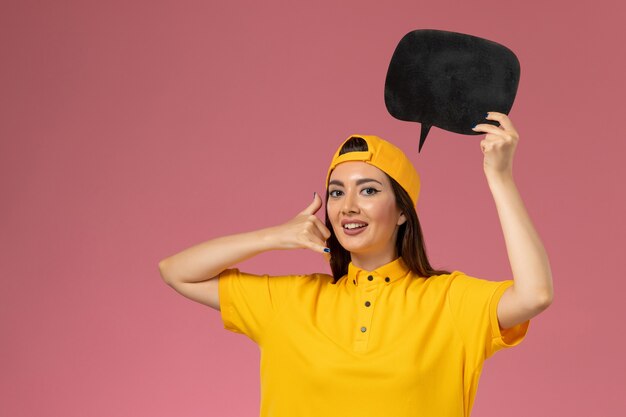 Front view female courier in yellow uniform and cape holding big black sign on the pink wall work service uniform delivery