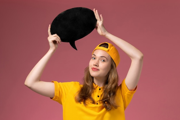 Corriere femminile di vista frontale in uniforme gialla e mantello che tiene segno nero sul lavoro dell'azienda dell'uniforme di consegna di servizio della parete rosa chiaro