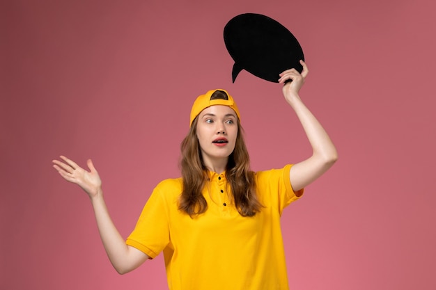 Corriere femminile di vista frontale in uniforme gialla e mantello che tiene segno nero sul lavoro uniforme di consegna di lavoro dell'azienda della parete rosa