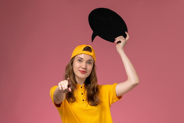 Front view female courier in yellow uniform and cape holding black sign on pink wall company service job delivery uniform work