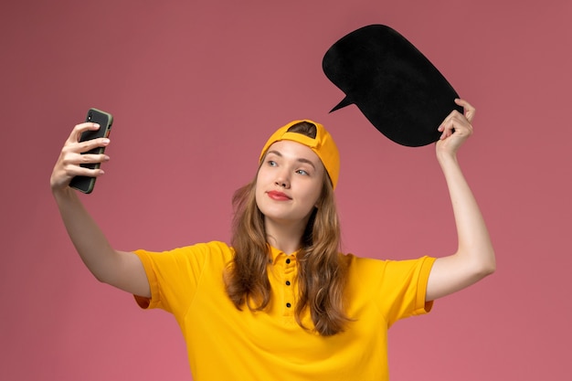 Front view female courier in yellow uniform and cape holding black sign and taking a photo on pink wall company service worker delivery uniform