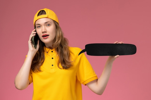 Corriere femminile di vista frontale in uniforme gialla e mantello che tiene segno nero e parlando al telefono sul lavoro di consegna di servizio dell'azienda uniforme della parete rosa