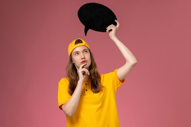 Front view female courier in yellow uniform and cape holding black sign and thinking on pink wall service delivery uniform