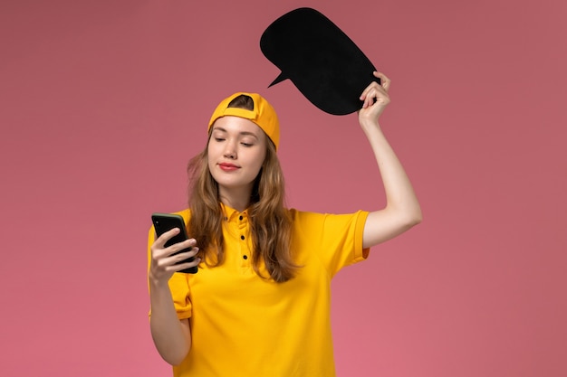 Corriere femminile di vista frontale in uniforme gialla e mantello che tiene segno nero e utilizzando il telefono sulla uniforme di consegna del lavoratore di servizio dell'azienda della parete rosa