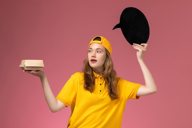 Front view female courier in yellow uniform and cape holding black sign with bowl on pink wall service delivery uniform company gitl worker