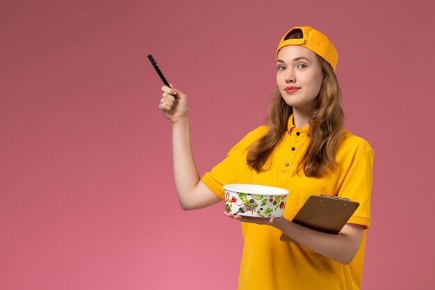 Вид спереди женщина-курьер в желтой униформе и накидке с миской для доставки и блокнотом с ручкой на светло-розовой стене.