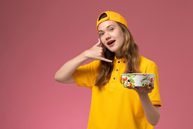 Front view female courier in yellow uniform and cape holding delivery bowl on pink wall service delivery job uniform girl