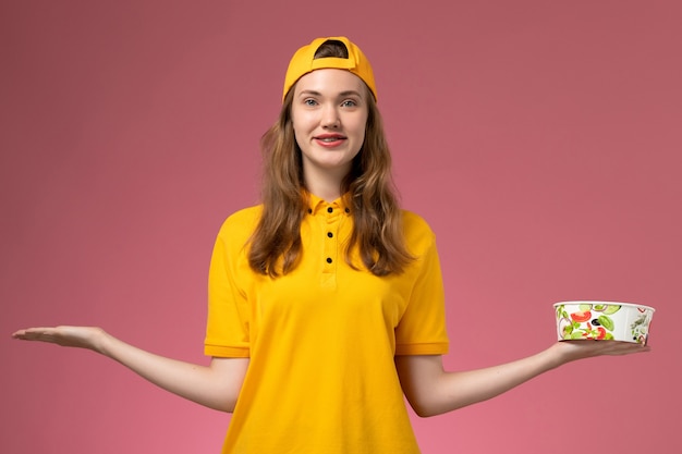 Вид спереди женщина-курьер в желтой форме и накидке, держащая миску для доставки на розовой стене, униформа для доставки услуг, работа компании