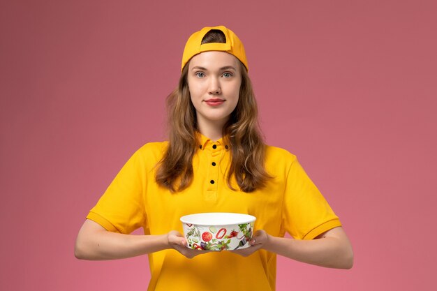 Front view female courier in yellow uniform and cape holding delivery bowl on pink wall service delivery uniform company job