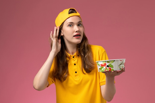 Front view female courier in yellow uniform and cape holding delivery bowl trying to hear on pink wall service delivery job uniform worker girl