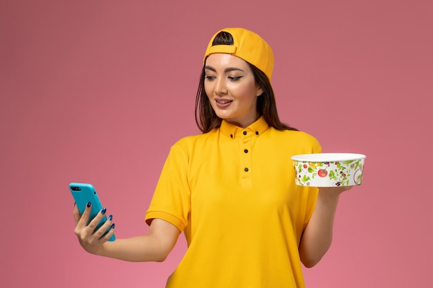 Front view female courier in yellow uniform and cape holding delivery bowl and using a phone on light pink desk company service uniform delivery job work