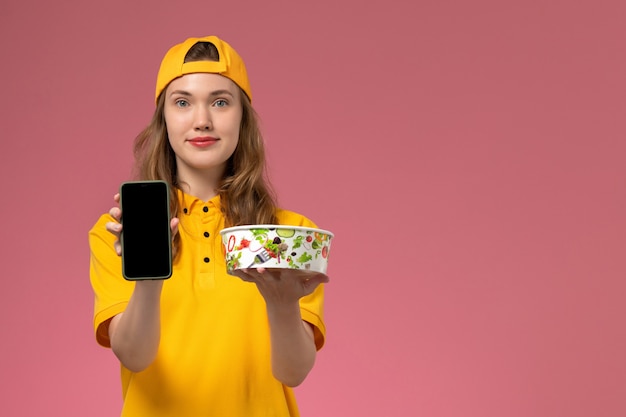 Вид спереди женщина-курьер в желтой униформе и накидке с миской для доставки с телефоном на розовой стене.