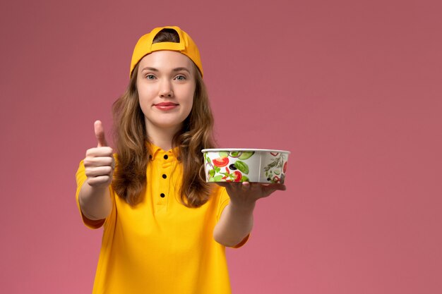 Front view female courier in yellow uniform and cape holding delivery bowls on pink wall service delivery job uniform work