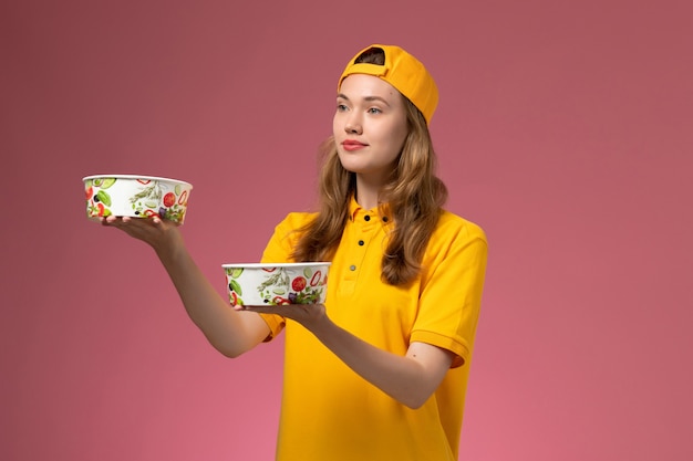 Front view female courier in yellow uniform and cape holding delivery bowls on pink wall service job delivery uniform girl worker