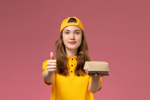 Front view female courier in yellow uniform and cape holding delivery food package on the pink wall service delivery job uniform company