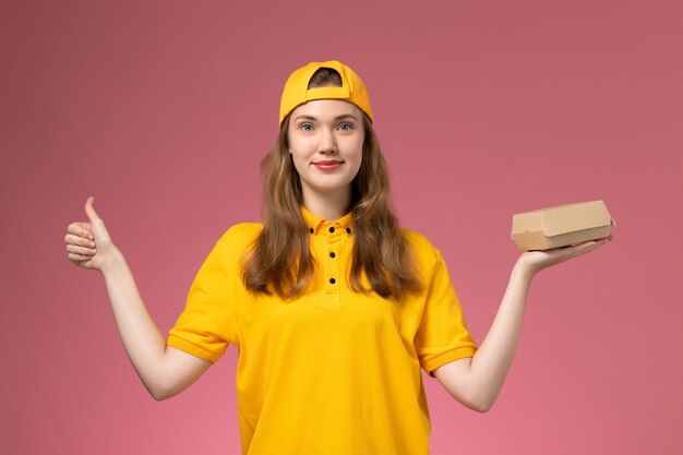 Front view female courier in yellow uniform and cape holding delivery food package on pink wall service delivery job uniform work