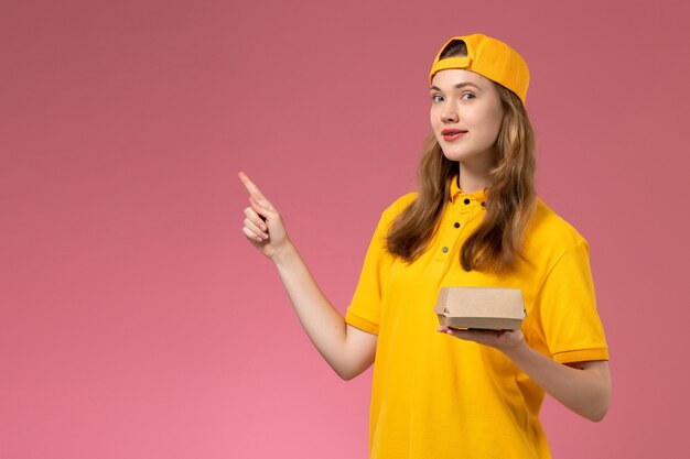 Front view female courier in yellow uniform and cape holding delivery food package on pink wall service delivery uniform company girl work