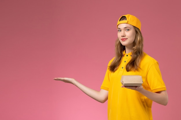 Corriere femminile di vista frontale in uniforme gialla e mantello che tiene il pacchetto di cibo di consegna sul lavoro di lavoro dell'azienda uniforme di consegna di servizio della parete rosa