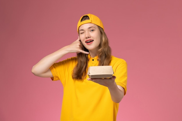 Front view female courier in yellow uniform and cape holding delivery food package on pink wall service delivery uniform company job worker