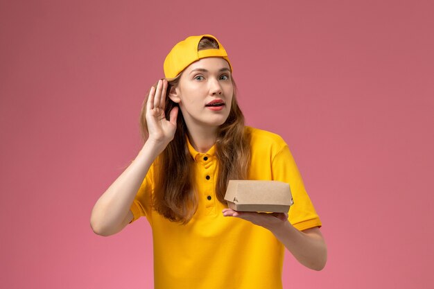 Front view female courier in yellow uniform and cape holding delivery food package on pink wall service delivery uniform company worker