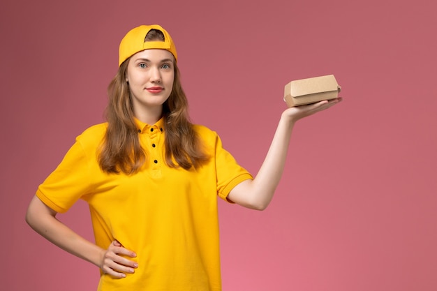 Front view female courier in yellow uniform and cape holding delivery food package on pink wall service delivery uniform job girl