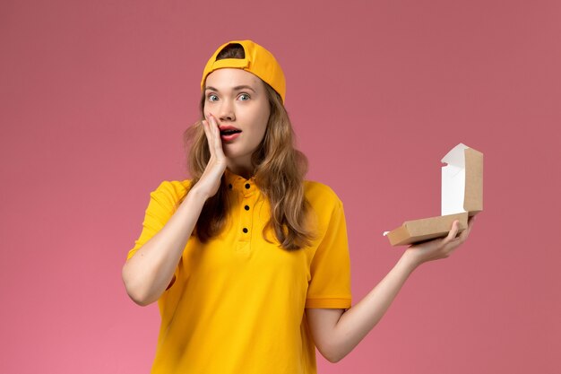 Front view female courier in yellow uniform and cape holding empty little delivery food package on light-pink wall job work service delivery uniform