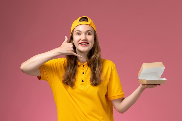 Front view female courier in yellow uniform and cape holding empty little delivery food package on light pink wall service delivery uniform